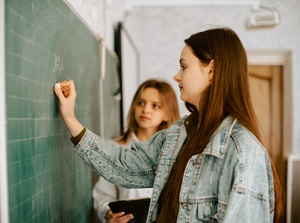 A person writes on a chalkboard while another looks on