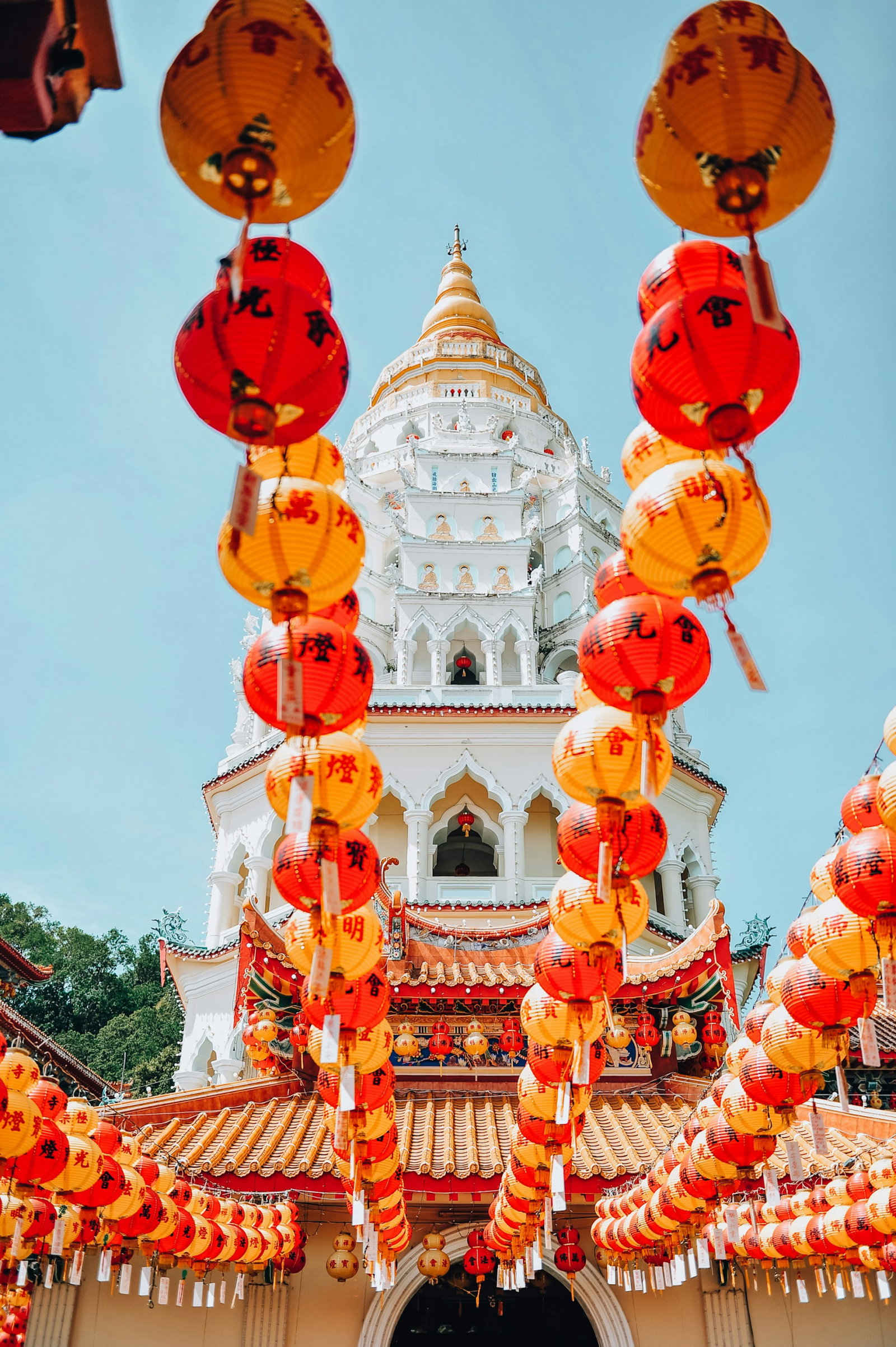 Red paper lanterns with inscriptions leading up to tiered white pagoda with orange accents and paintings of the Buddha