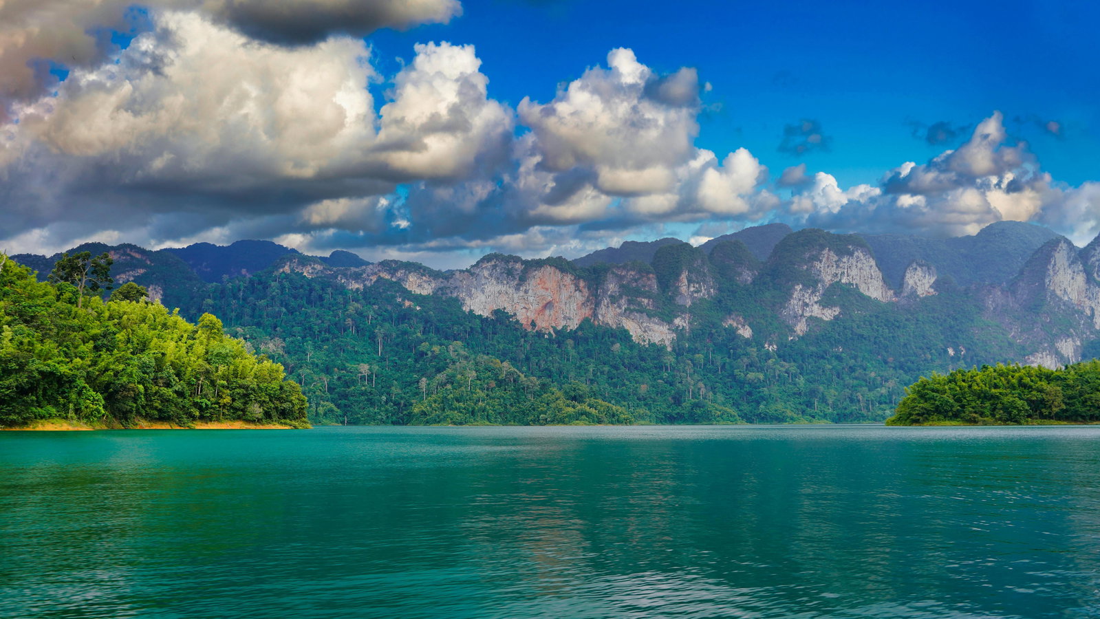 Dark green lake with lush island to the left and tall mountains in the centre, grey clouds in bright blue sky