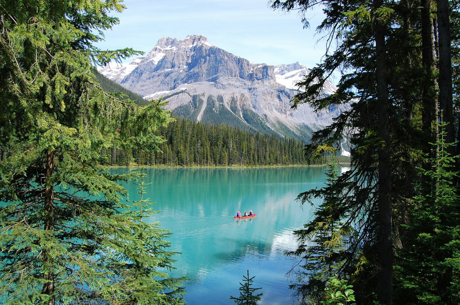 Nature viewpoint with pine trees and bright turquoise lake with canoe boat in the centre, backed by grey mountains