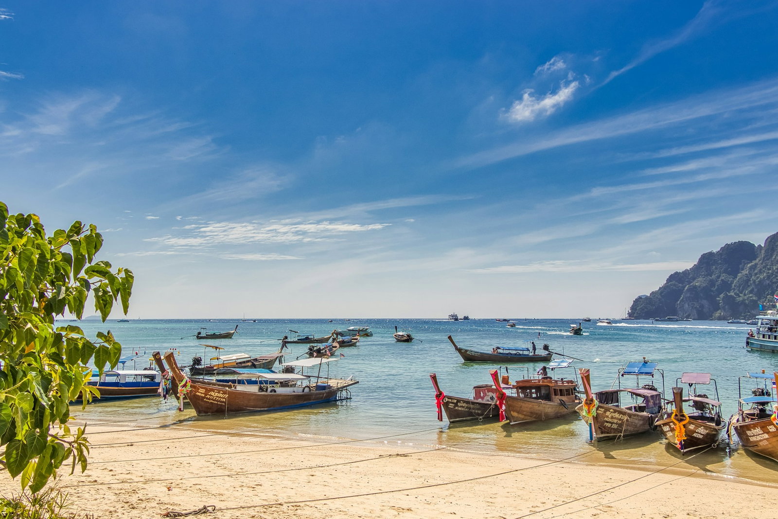 Golden sandy beach with traditional longtail boats tied to the shore, more boats in the water on a blue sky day