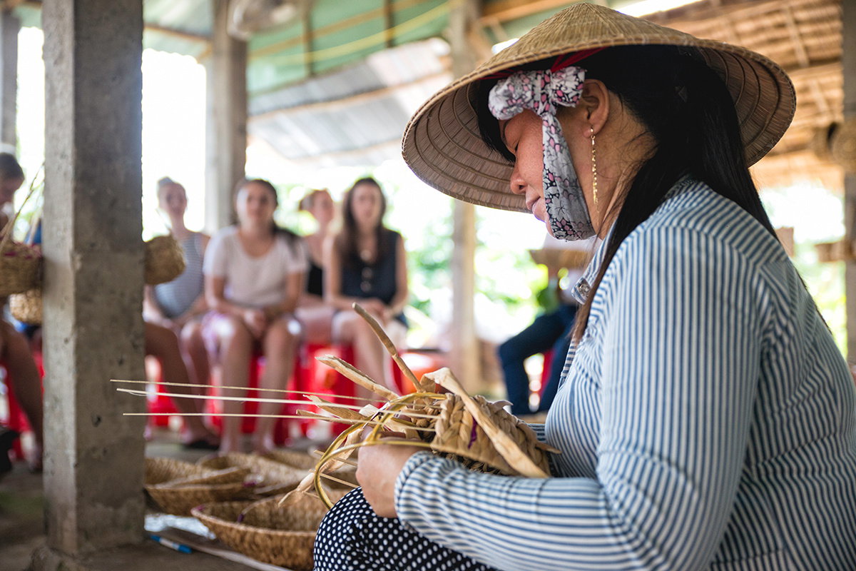 A lady weaves a straw hat while a group of travellers watch
