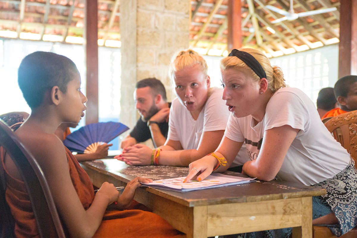 Two volunteers helping a child in monk robes read a book
