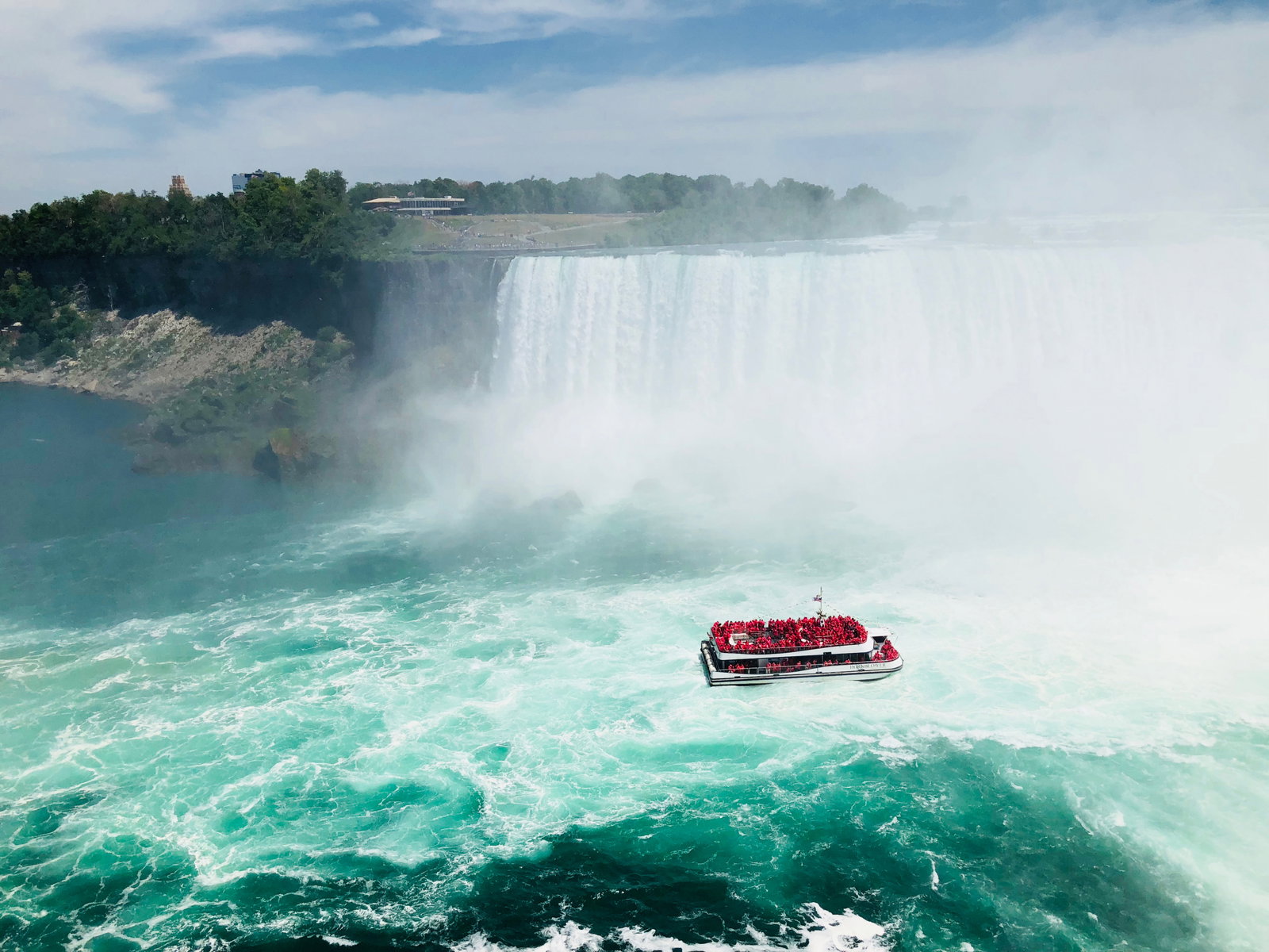 Boat floating through the waters of epic waterfall surrounded by green shrubbery and some modern city buildings