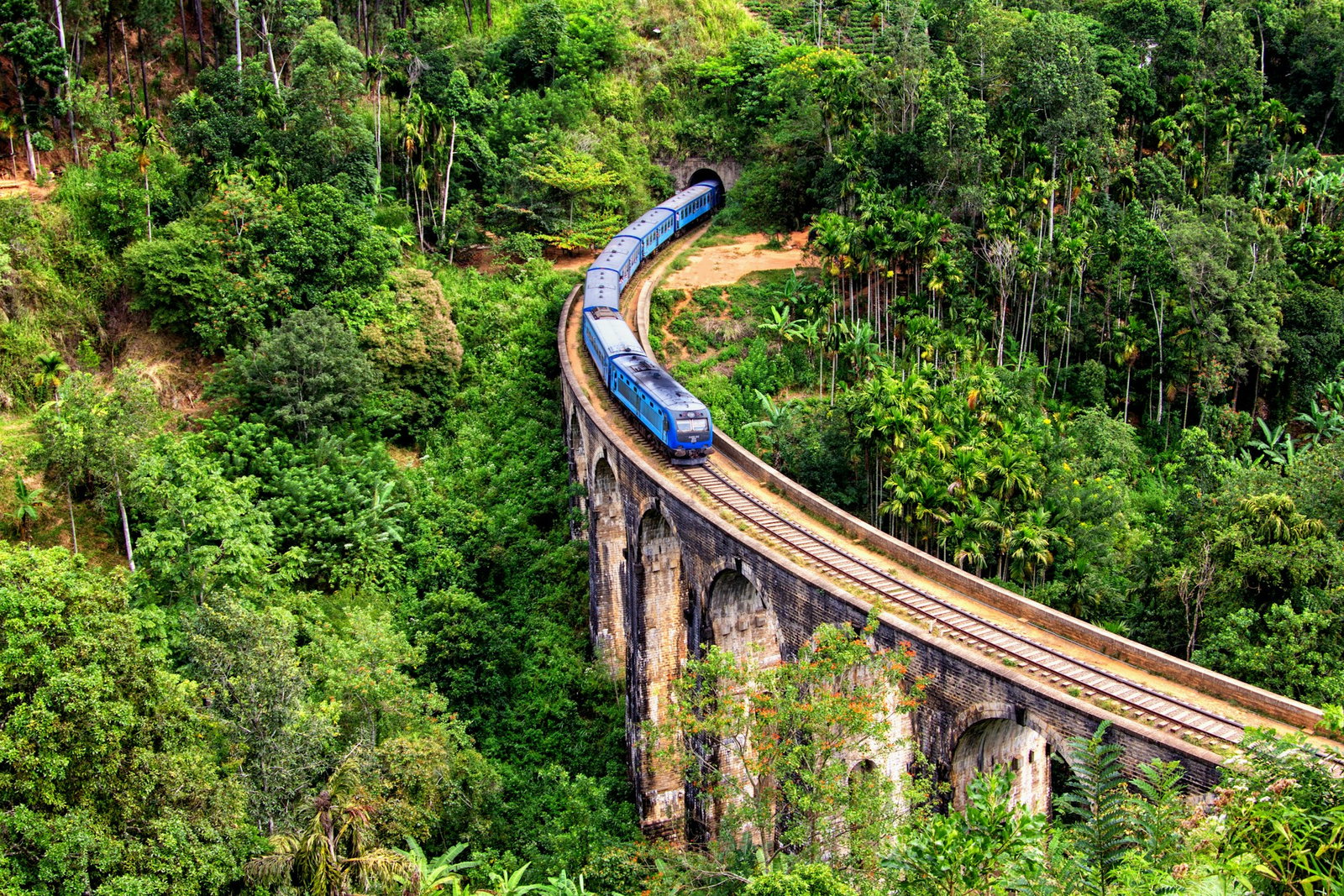 Aerial shot of emerald forest and old stone bridge with blue train gliding along the tracks