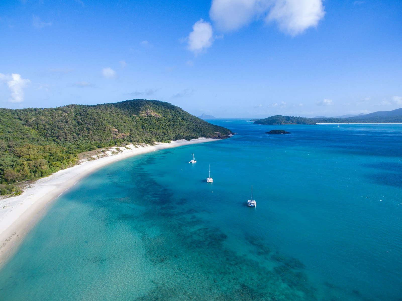 Blue ocean with three sailboats, curved white sand and lush forest, little islands in distance, sunny day with clouds