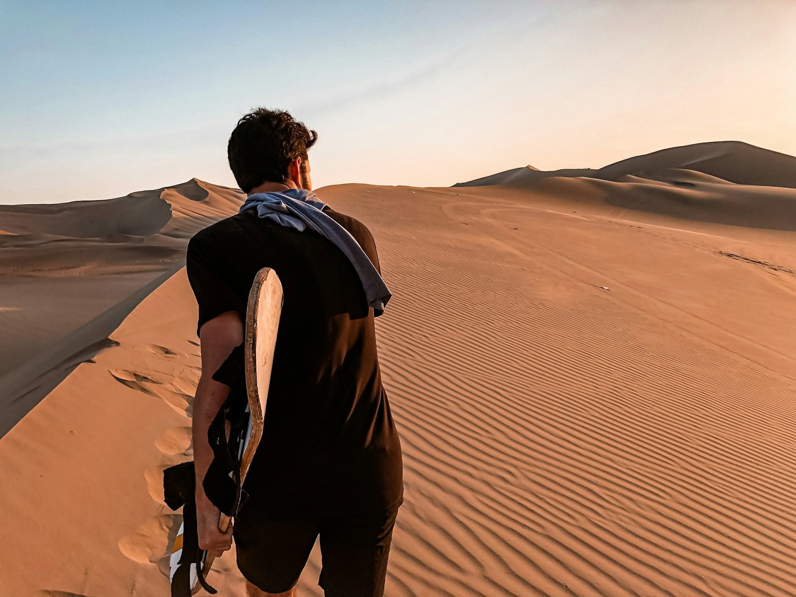 Person in black clothing carrying a sled and walking over golden dunes in the desert at sunset, Huacachina