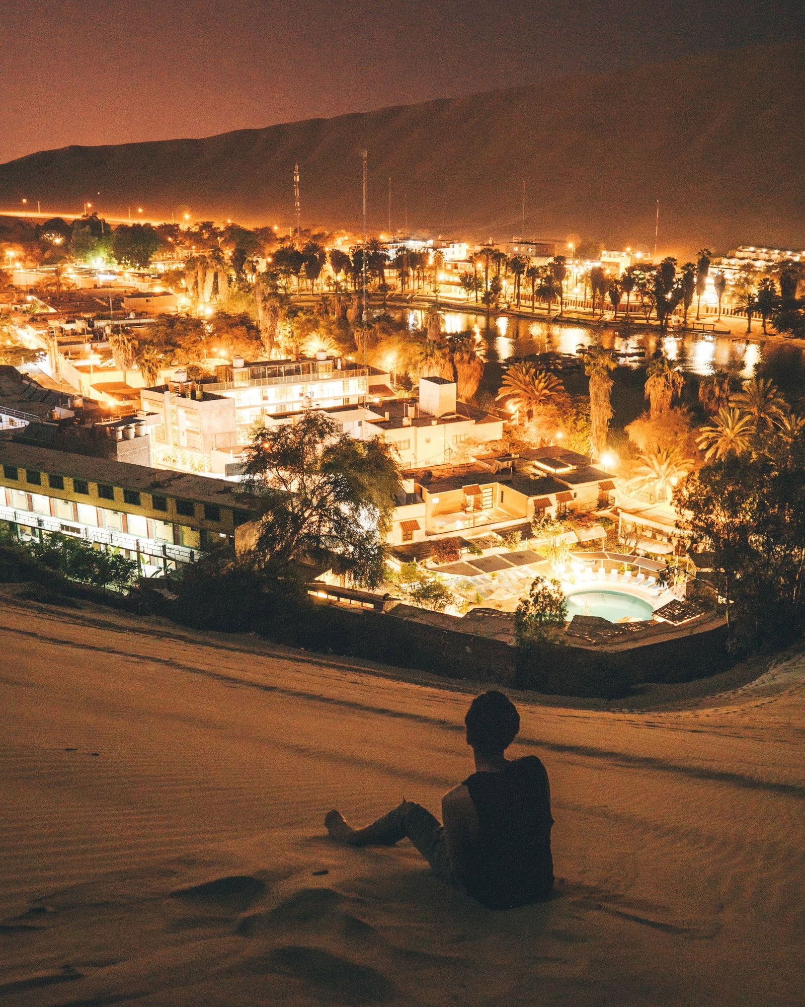 Person sitting on desert dune overlooking city oasis in the centre illuminated by golden lights at night, Huacachina