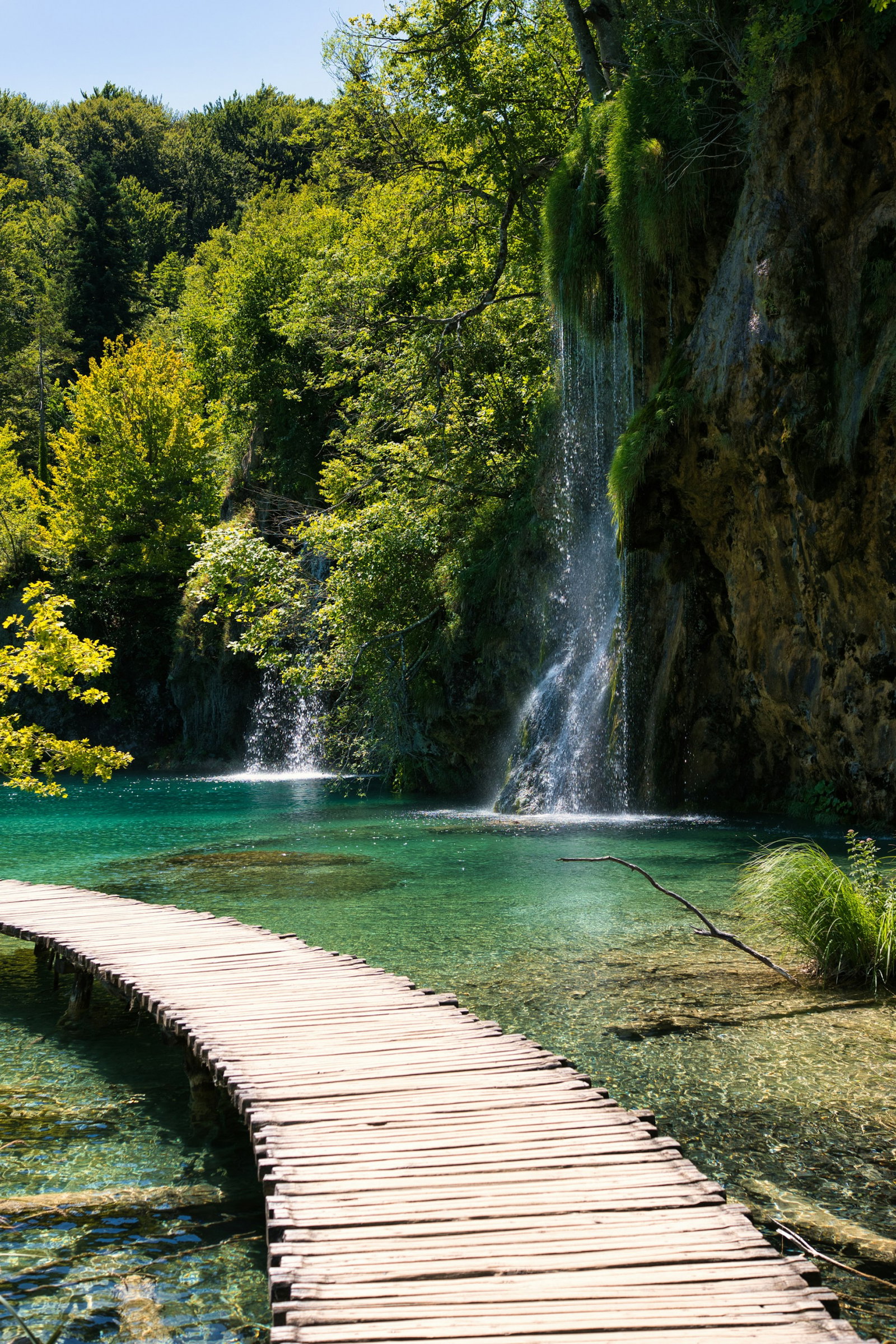 Wooden boardwalk across bright turquoise lake with green forest and thin, towering waterfall