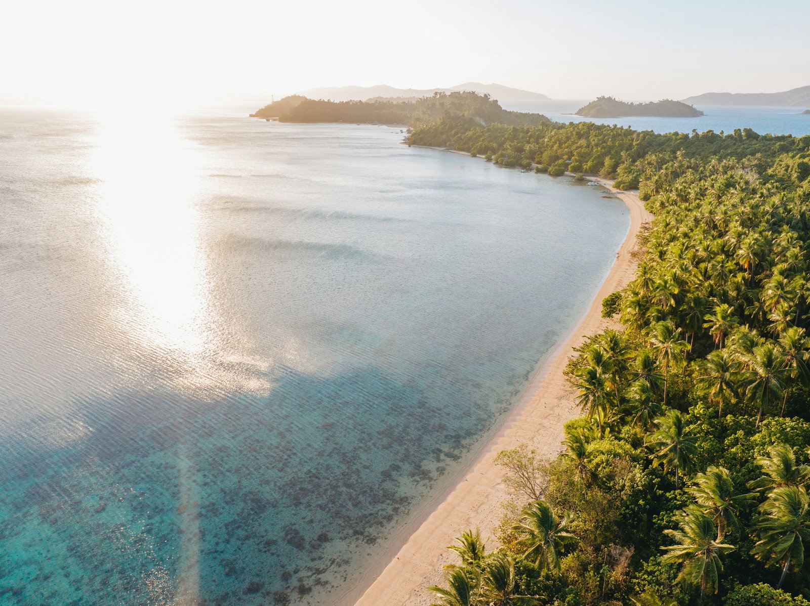 Sun shining on blue ocean with long stretch of golden sand and lots of palm trees on island with little bays in the distance