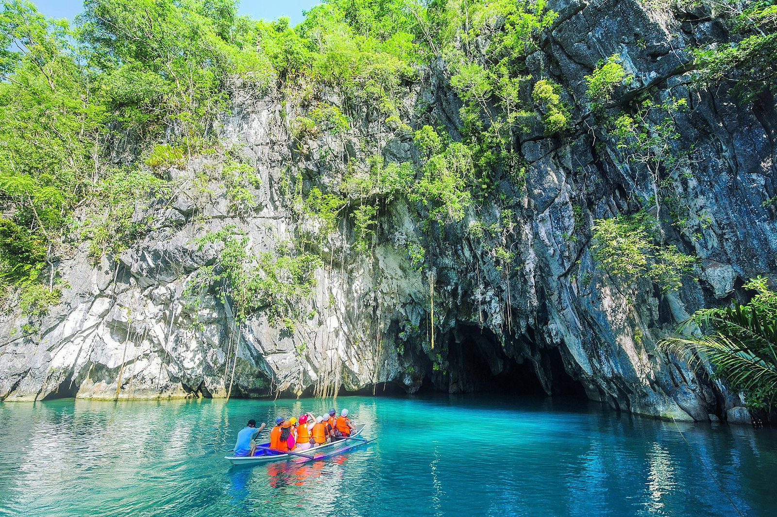 Little boat gliding over bright sapphire waters into a grey cave covered in emerald jungle