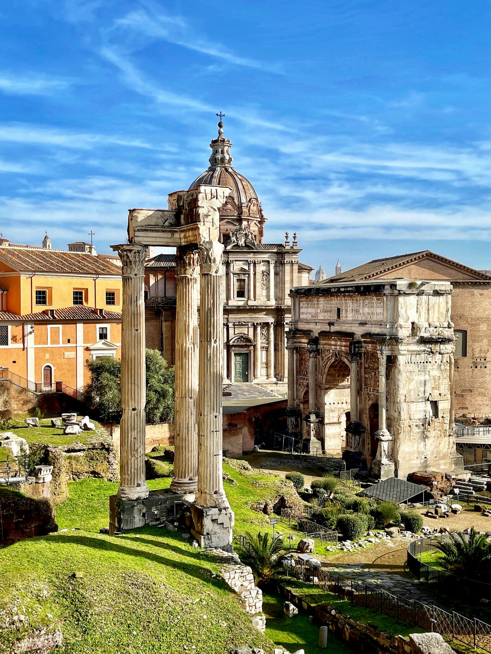 Ancient Roman ruins and columns on green hilltops surrounded by more modern colourful buildings on a clear blue sky day