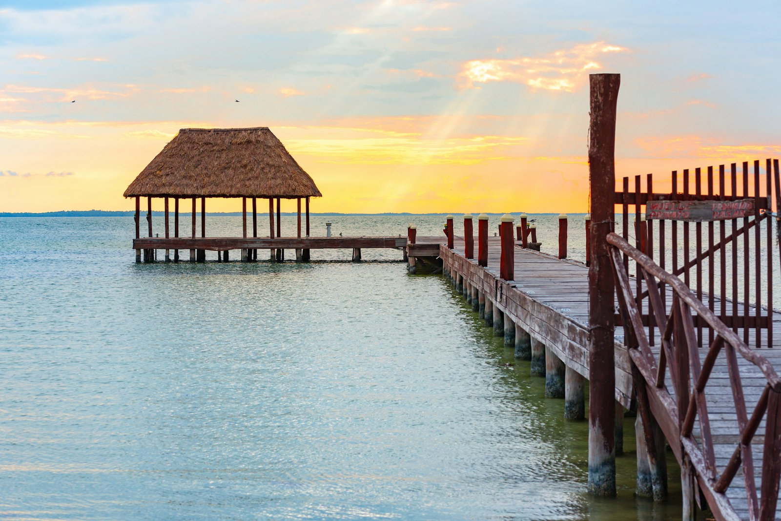 Rustic boardwalk and pier with thatched roof on glistening blue ocean at sunset with pale oranges and blues