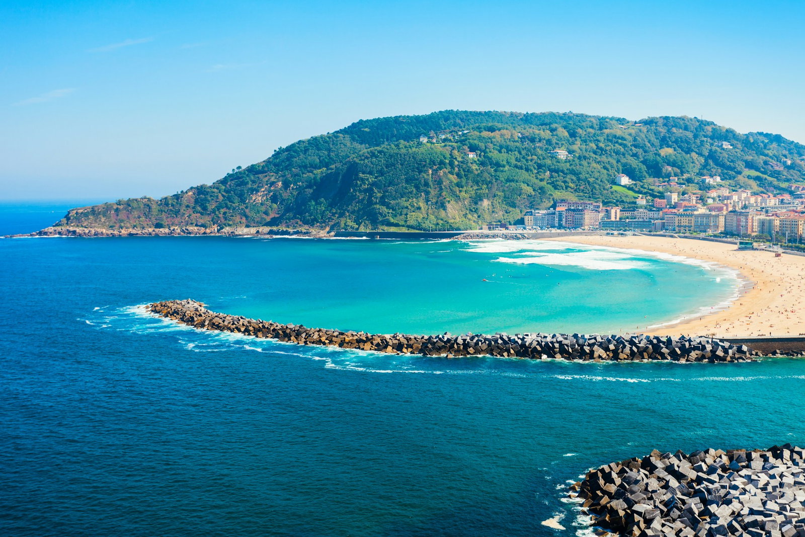 Bright sunny day with blue ocean, golden sand and rocky boardwalk with green hills and city buildings in backdrop