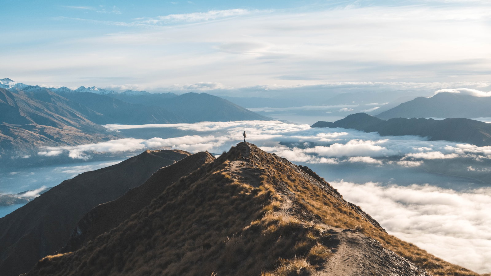 A lone traveller stands on top of a mountain with a blanket of clouds below