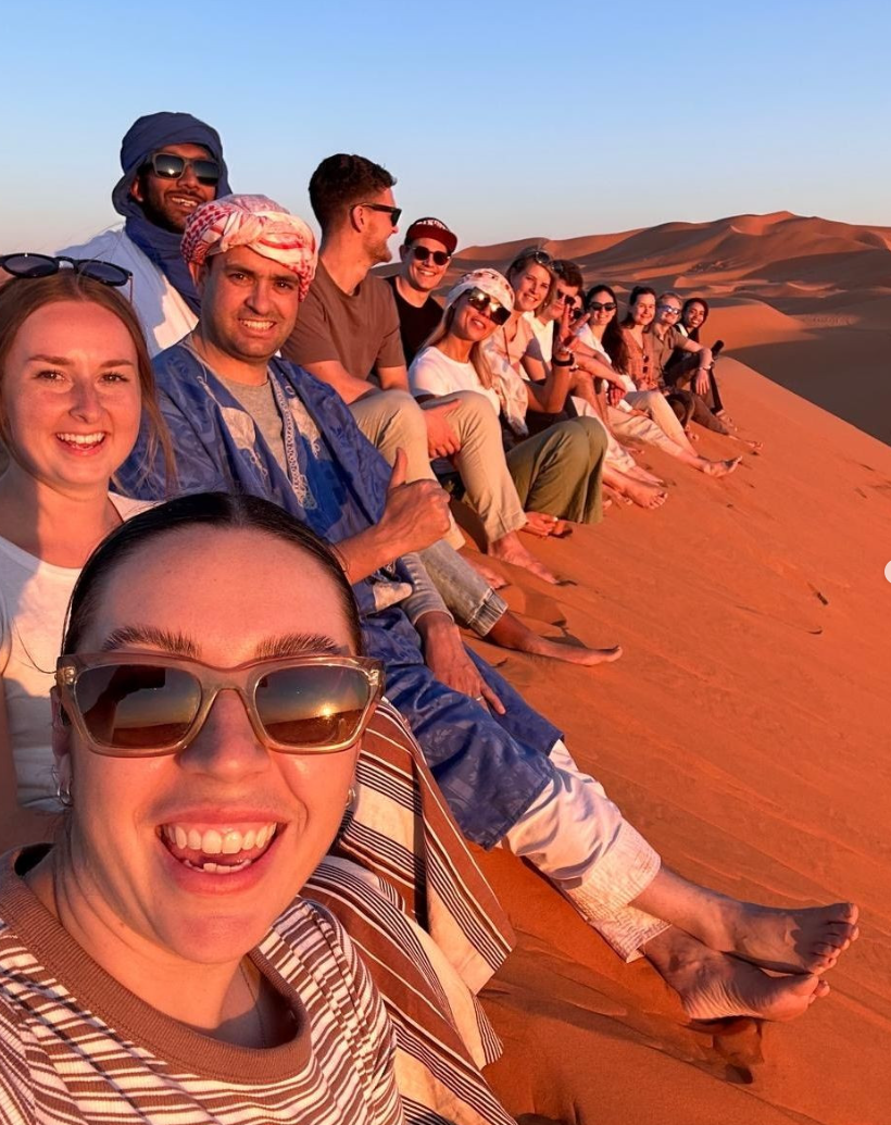 Group of travellers sat at the top of a sand dune in the Sahara Desert at sunset in Morocco