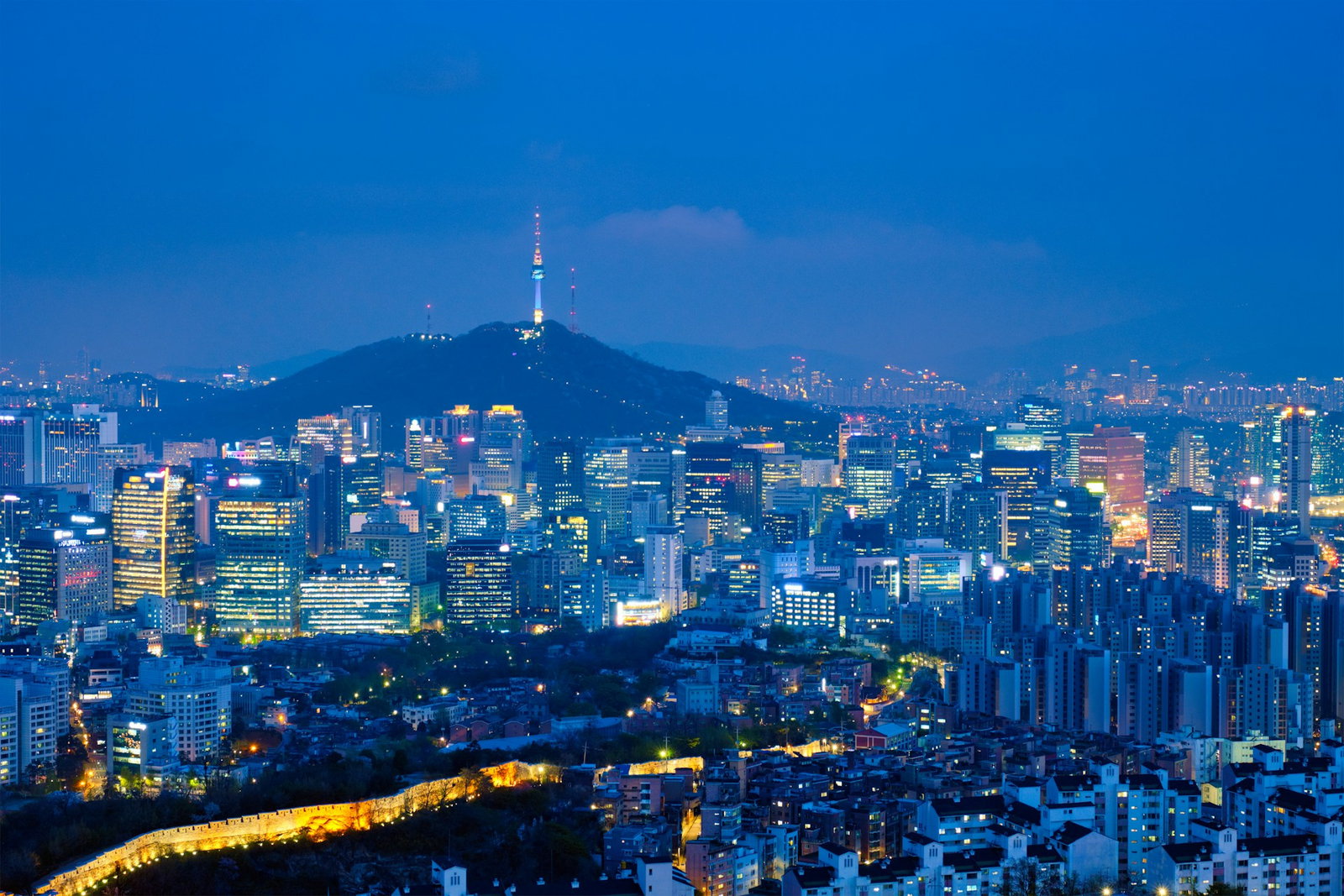 Modern cityscape at night with skyscraper blocks, traditional ancient walls and forested hills with tower at top