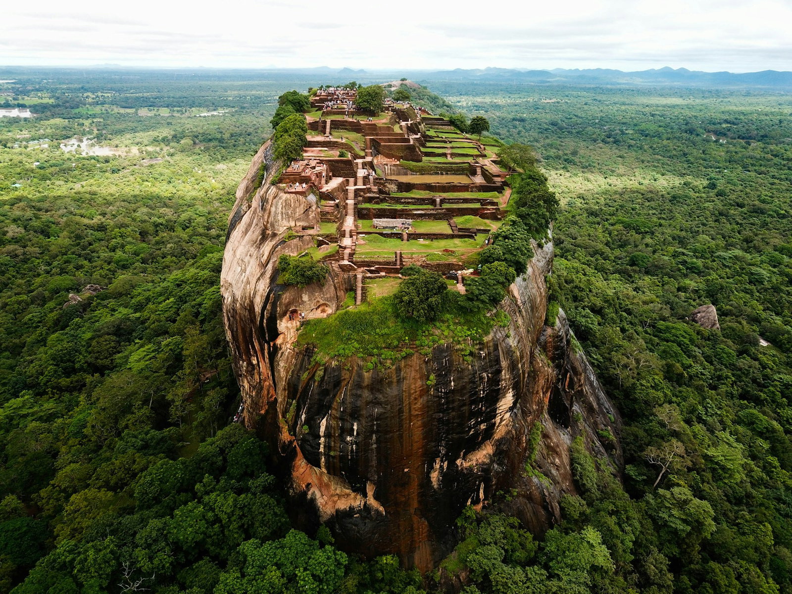 Aerial shot of giant monolith rock looking over a forest with a complex of ruins at the top