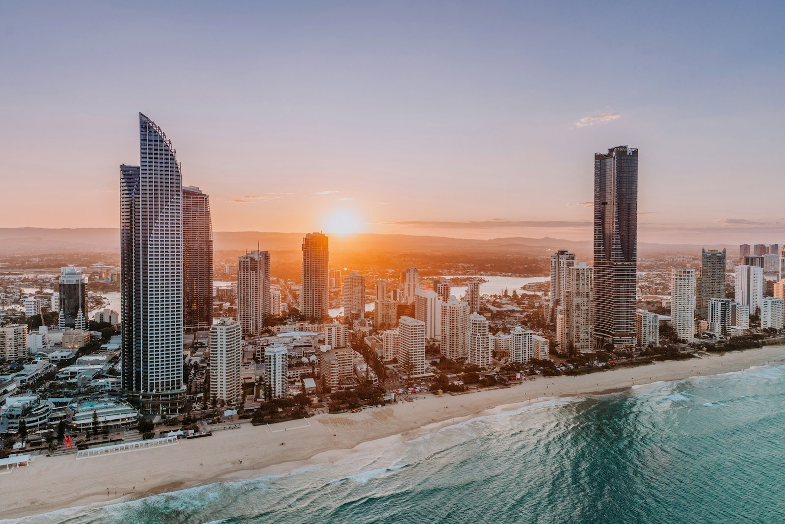 Modern skyscrapers by stretches of white sand and ocean at sunset, lake in the distance, Australia