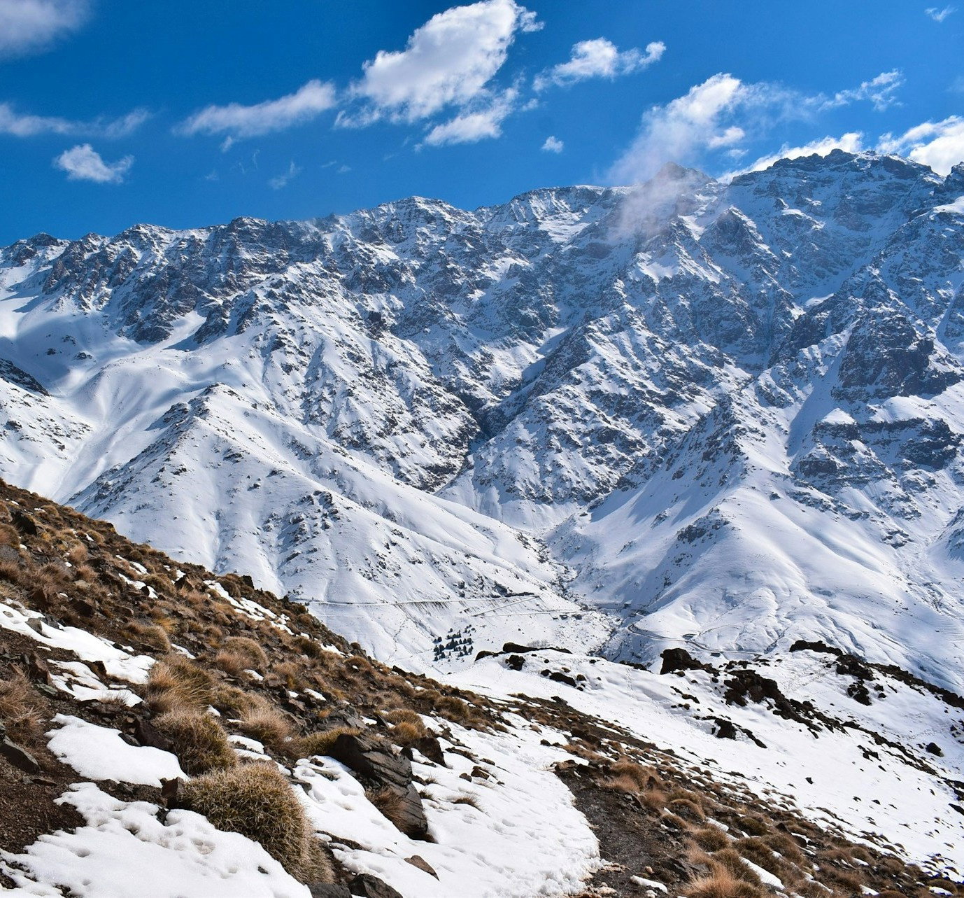 Rugged mountains covered in snow with some grassy terrain and blue skies with some clouds