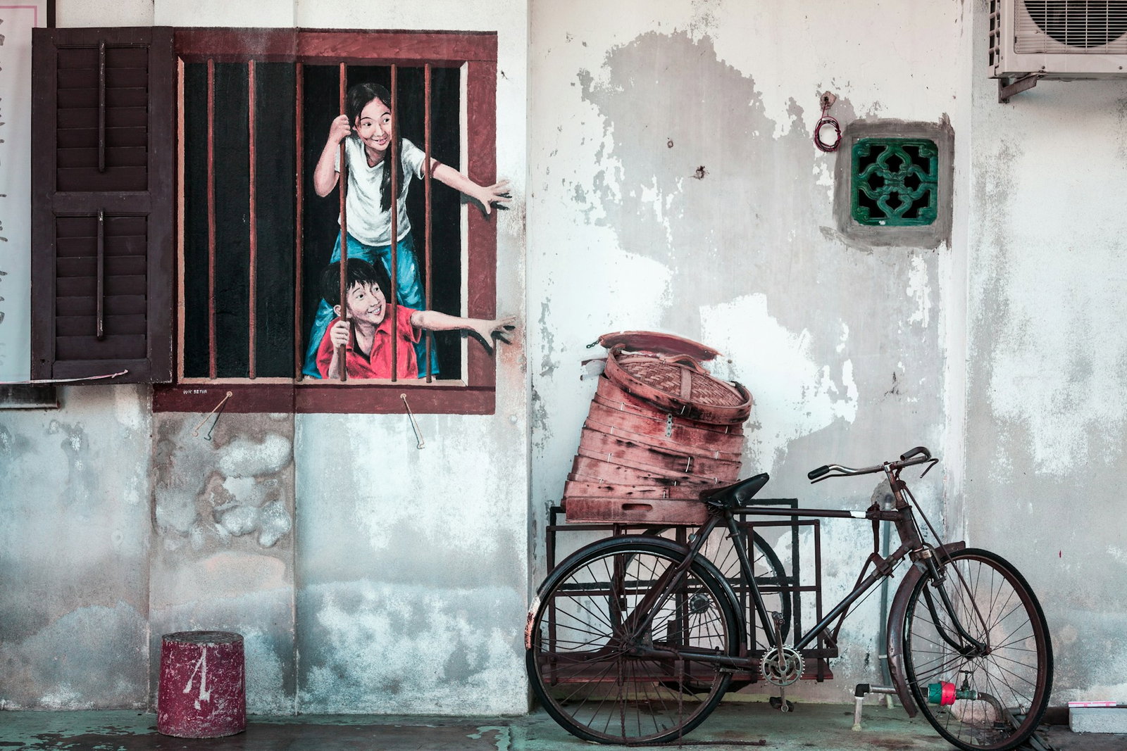 Street art mural depicting 2 children smiling and reaching out of window bars to a real bike with a basket on it