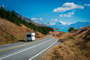 Mobile home driving along long road in New Zealand surrounded by dry grass, forest and blue mountains with lake
