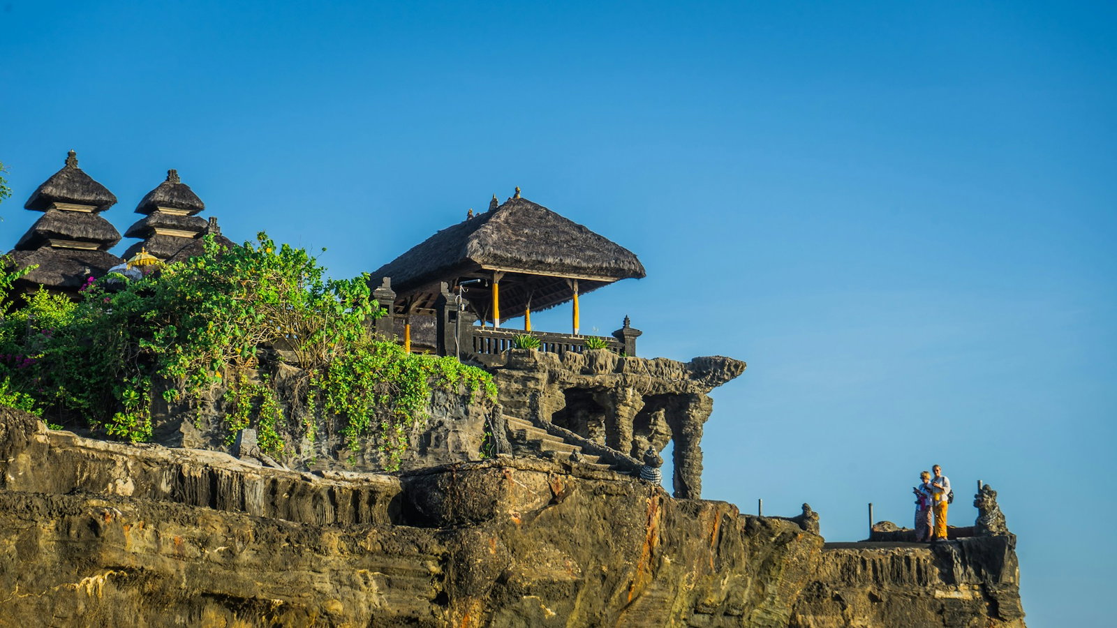 Traditional Balinese temple with thatched grey roofs sitting atop of rugged cliffside on a clear blue day, Tanah Lot Temple