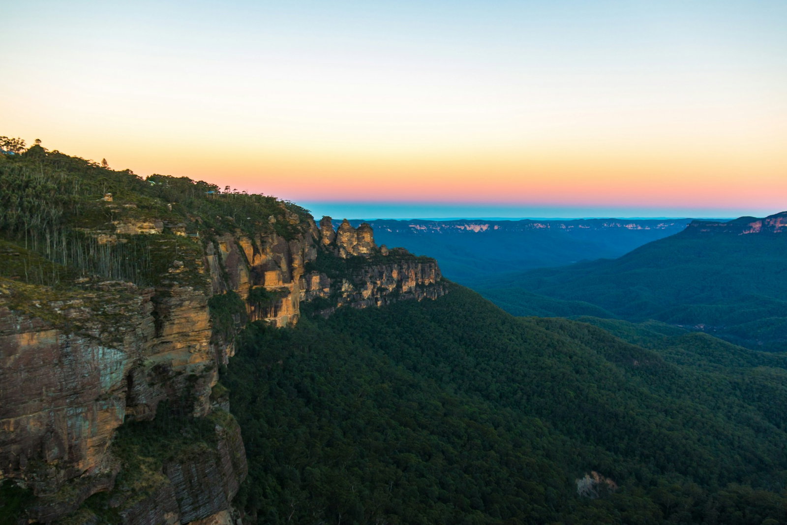 Rugged cliffs leading into hills covered in dense dark forest under a sunset sky, The Blue Mountains