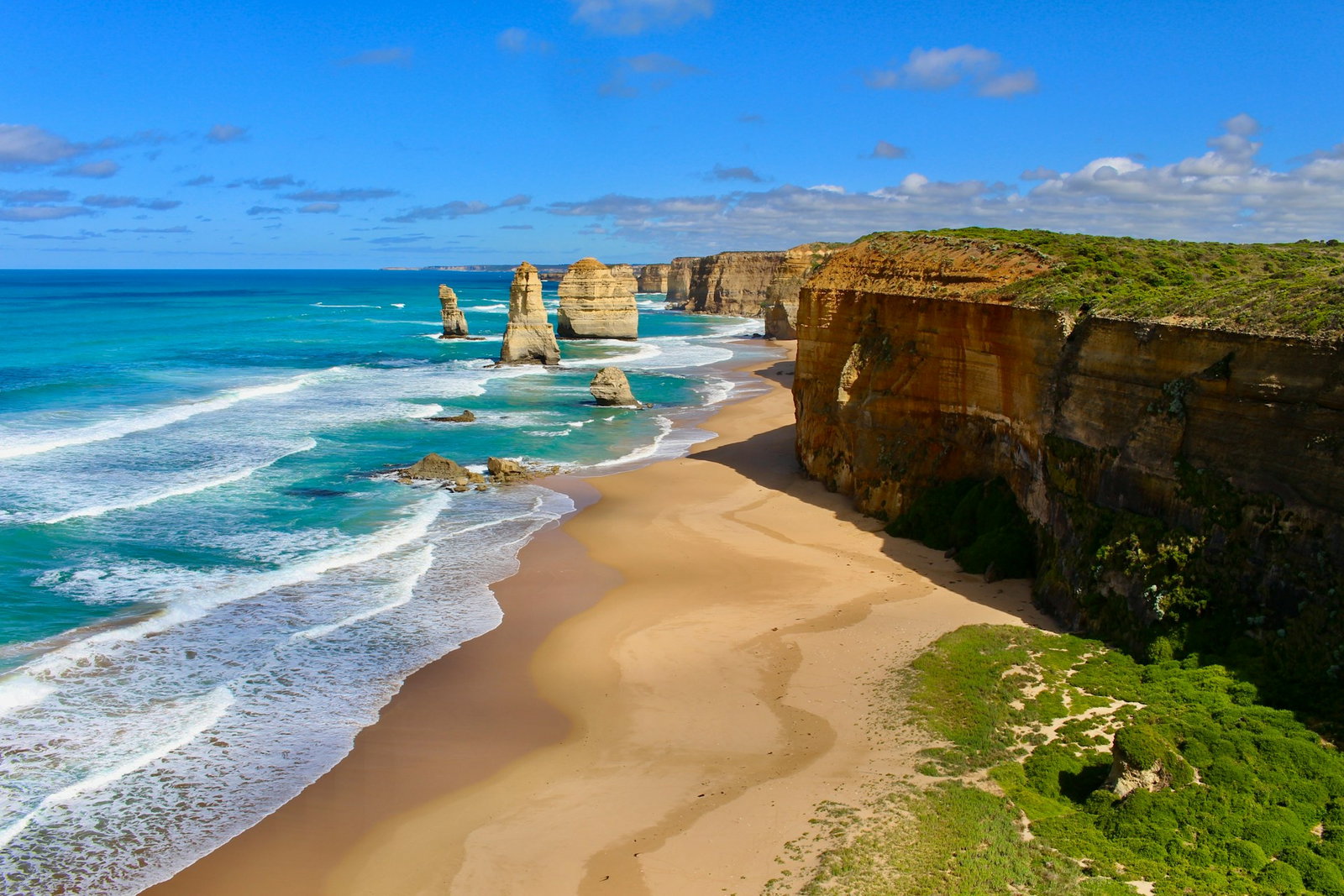 Gold sand with bright blue waves lapping at the shore, mossy brown cliffs and rock stacks in the ocean