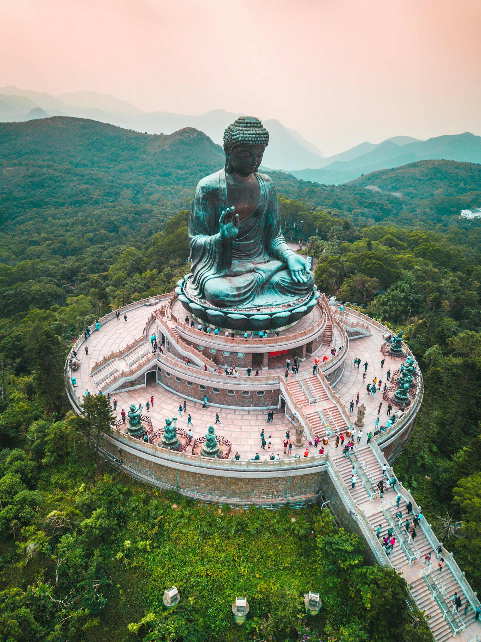 Giant blue statue of the Buddha sat on a rounded tower with stairs and tourists in the mountains