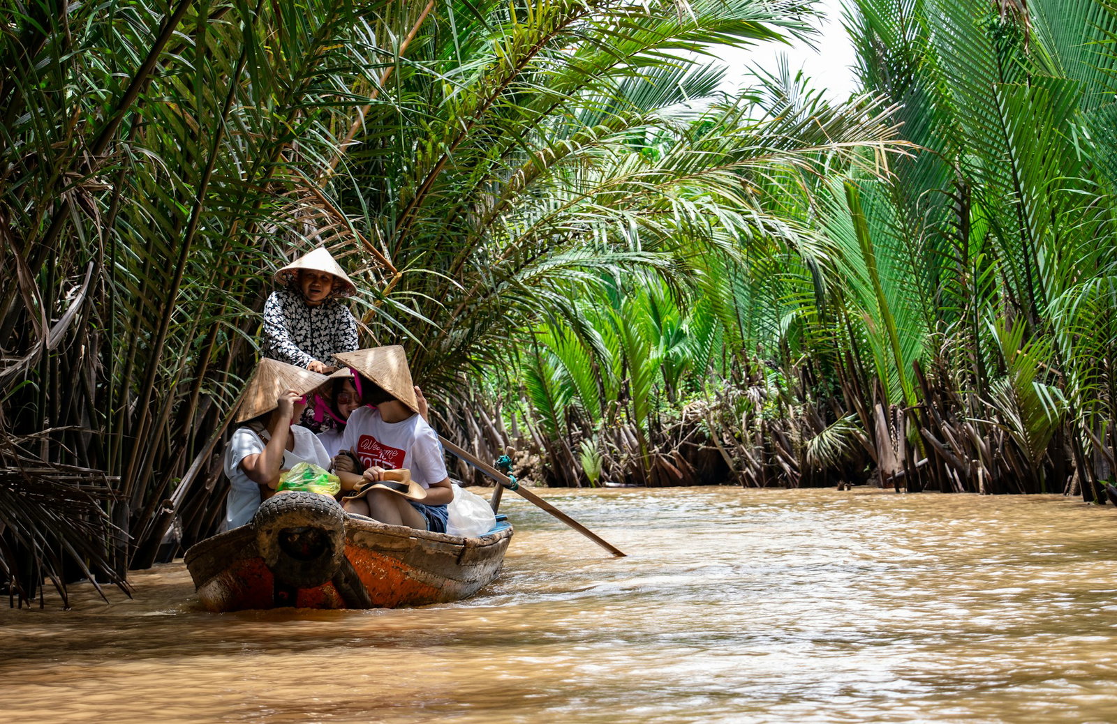 Travellers wearing strawhats travelling down a waterway lined with palm trees in the Mekong Delta in Vietnam