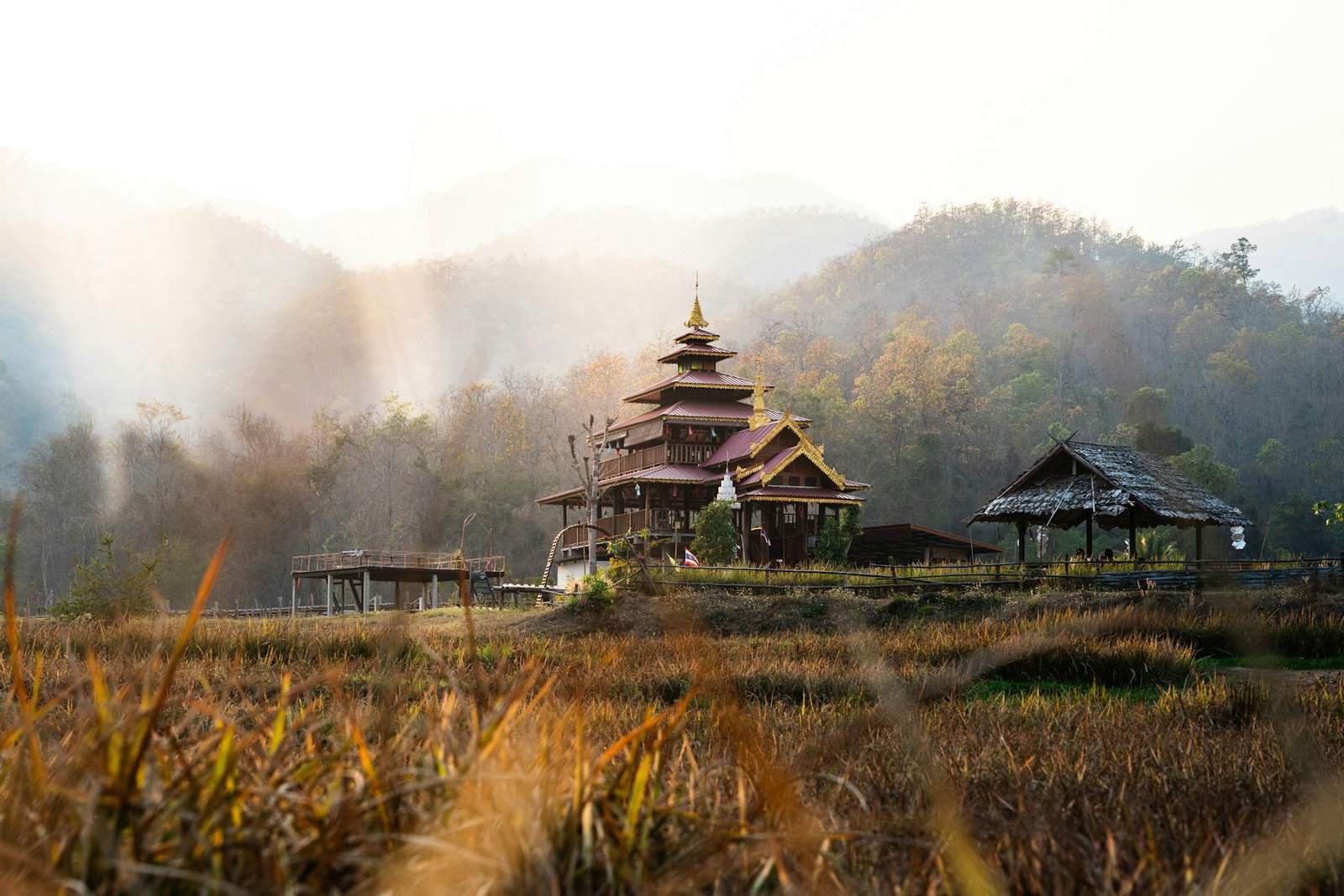 Traditional pagoda-style house with rustic wooden bridges in distance, surrounded by dry grass fields and forested hills