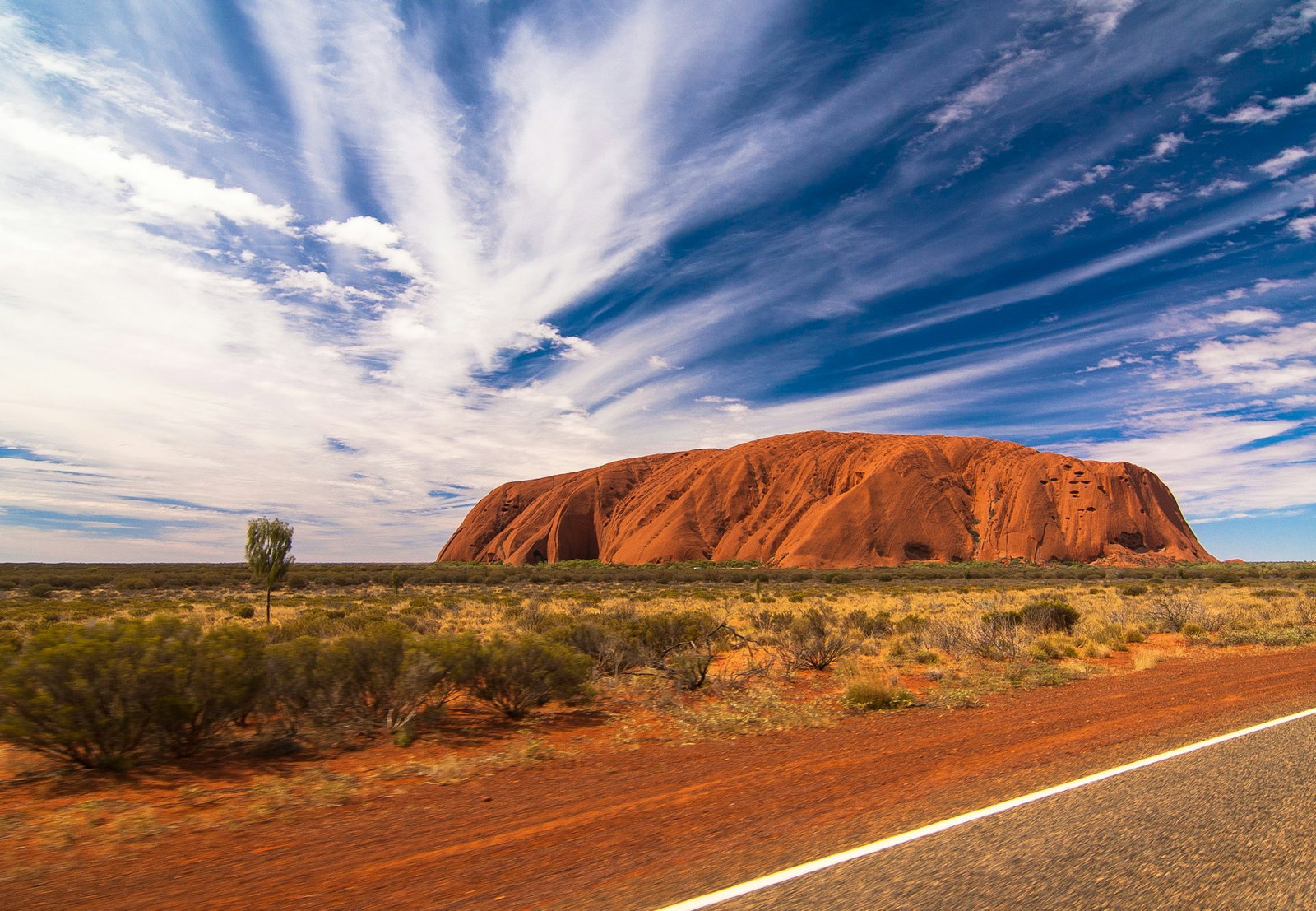 Giant red monolith rock surrounded by dry green and yellow desert on side of road, cloudy blue sky, Uluru