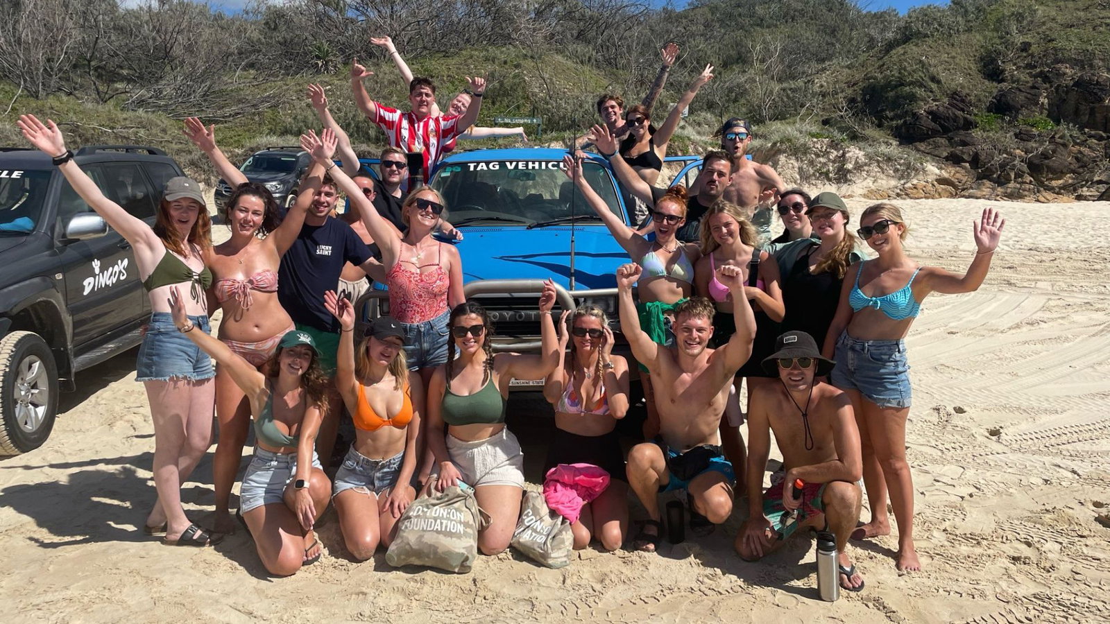 Group of travellers in swimsuit on sandy beach smiling and waving in front of blue vehicle