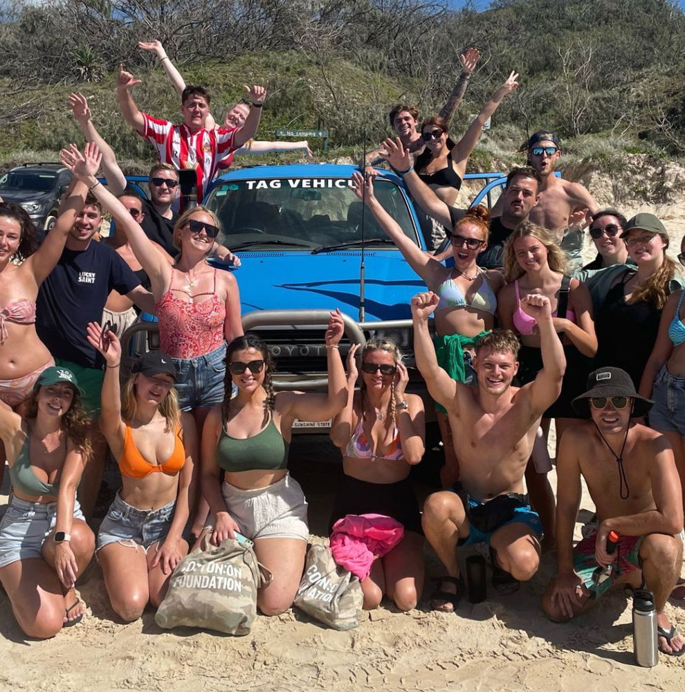 Group of travellers in swimsuit on sandy beach smiling and waving in front of blue vehicle