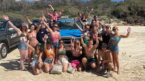 Group of travellers in swimsuit on sandy beach smiling and waving in front of blue vehicle
