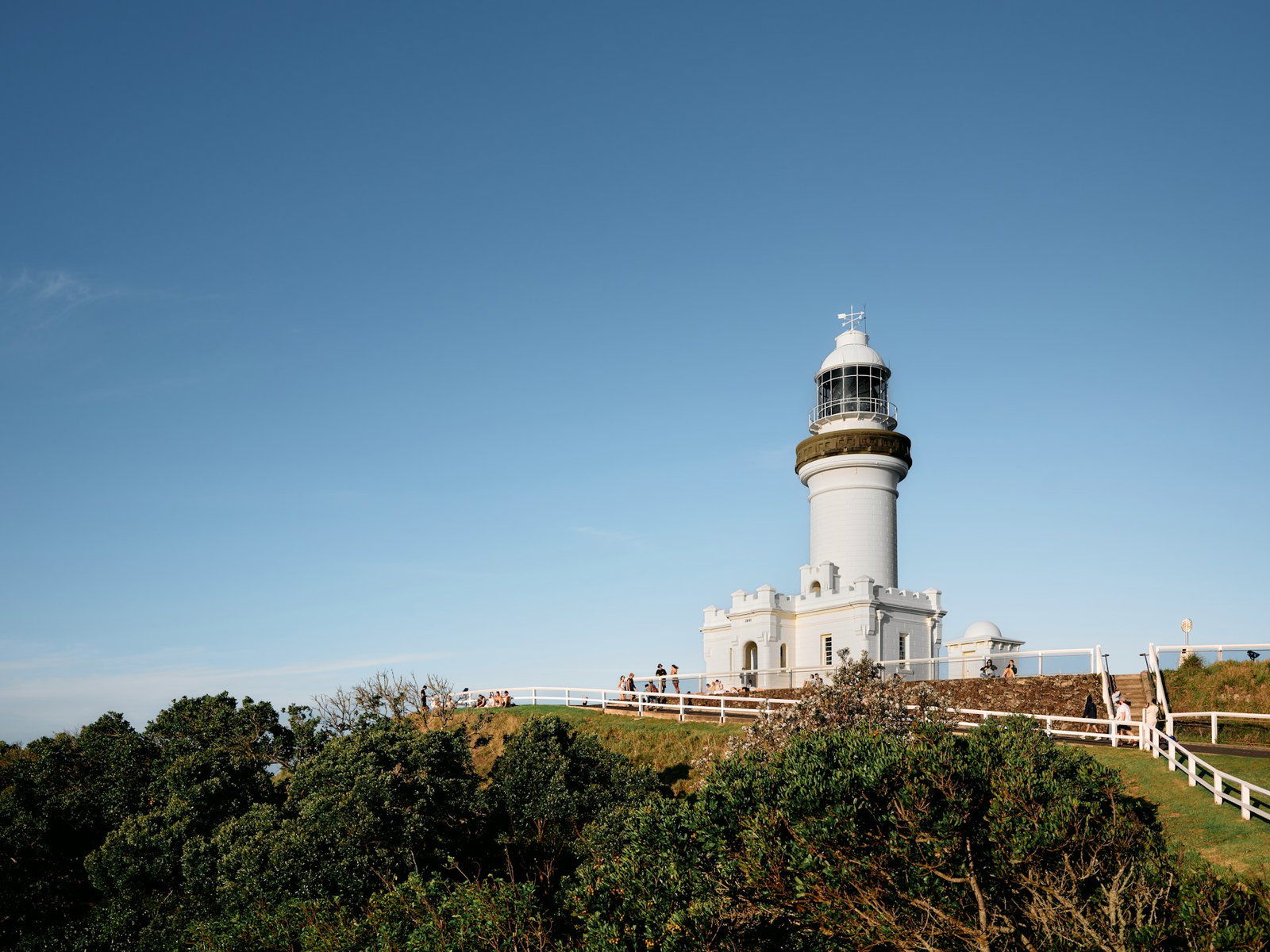 White lighthouse on green hill with people admiring the view on sunny day, Byron Bay