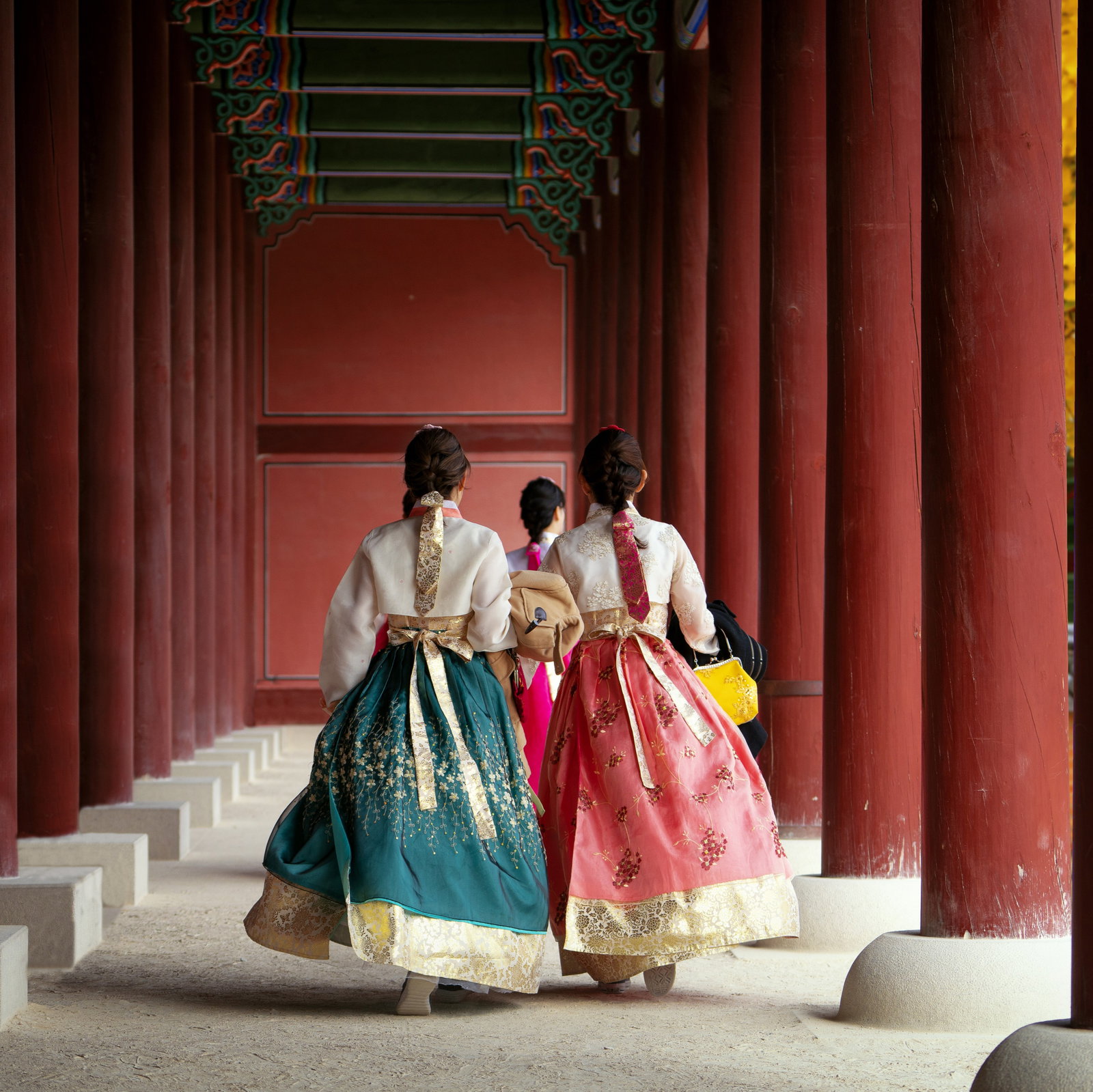 Three women dressed in traditional and colourful hanbok dresses and carrying bags as walk through maroon palace columns