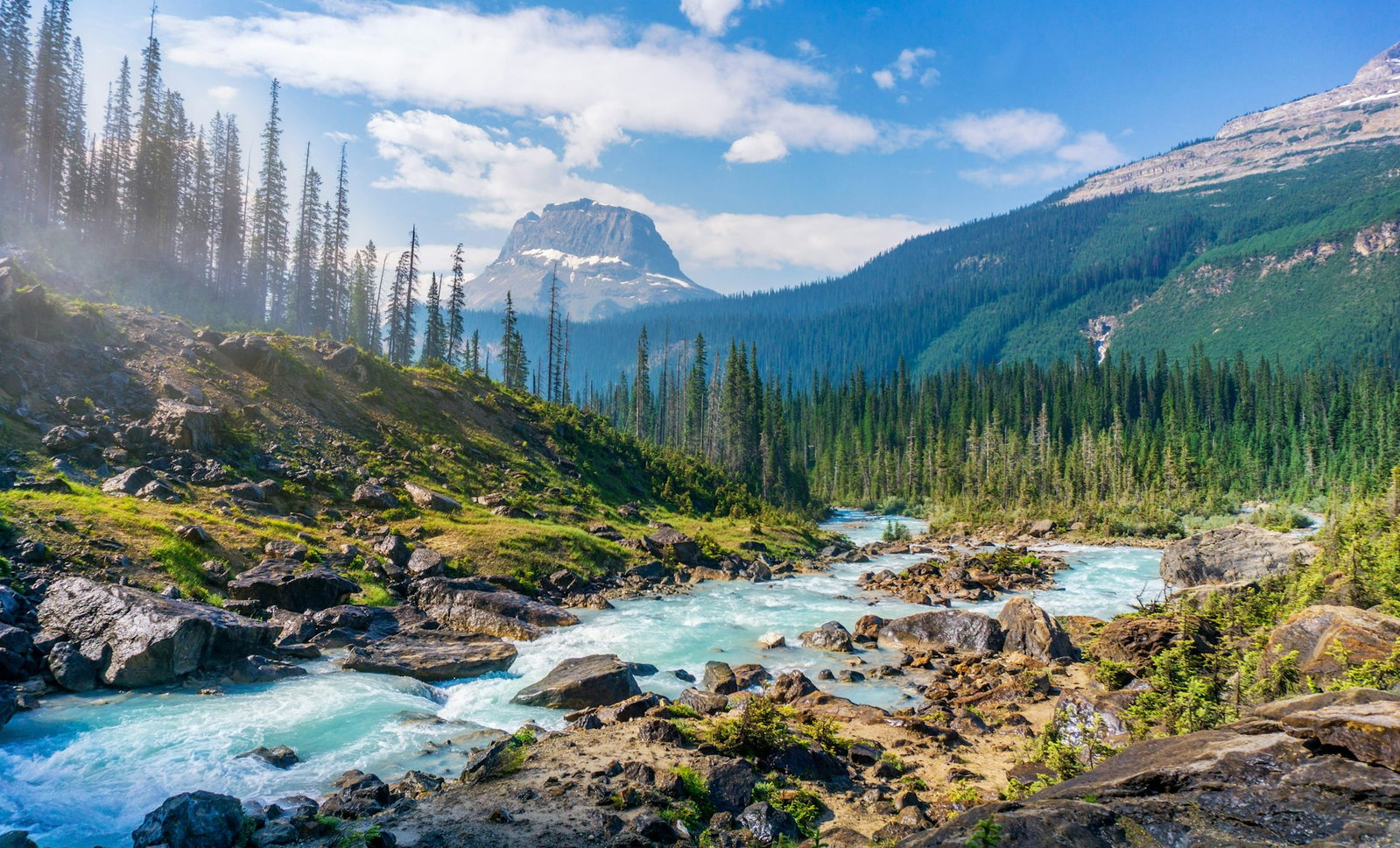 Quiet river stream with rocks and sloped green hills surrounded by pine forest with rugged grey mountain and blue cloudy sky